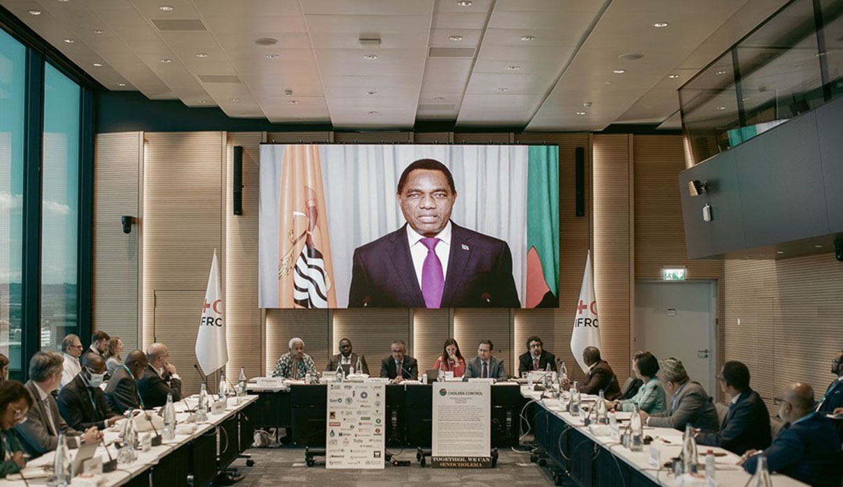 Attendees from different countries sitting in a conference room listening to a presentation on a video screen.
