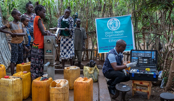 A group of adults and children in a jungle looking at a WHO doctor taking out medical equipment from a box.