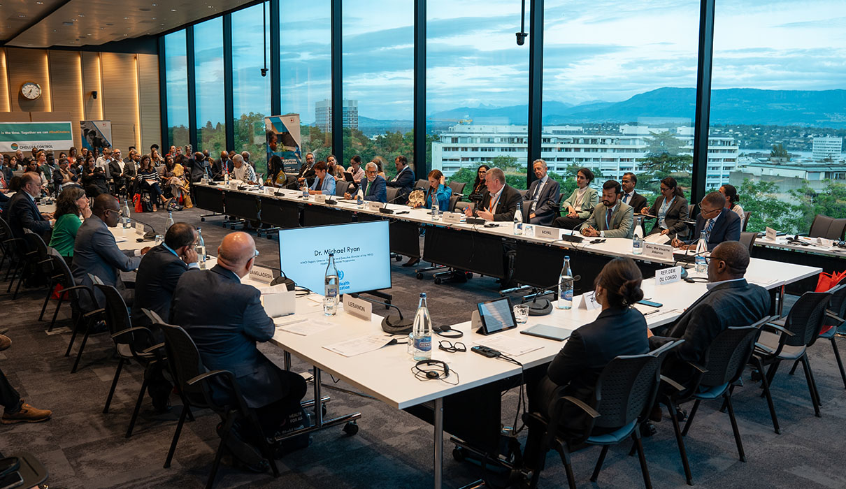Attendees from different countries sitting in a rectangle in a conference room listening to a presentation on a video screen.