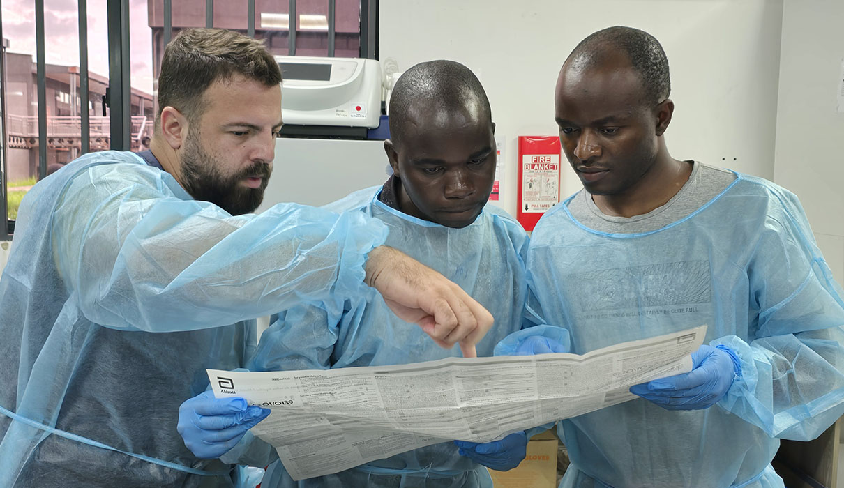 3 lab technicians wearing prtotective clothing looking at a laboratory manual.